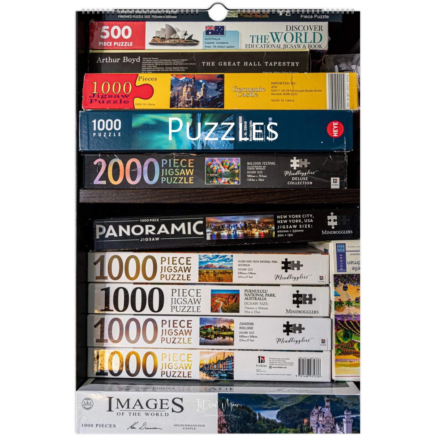 A tall retail display or shelf filled with boxed jigsaw puzzles from multiple brands, including Arthur Boyd, The Great Hall Tapestry, Woodland, Panoramic, and Images of the World. The boxes are stacked horizontally, showing different puzzle types and piece counts. 2027 Puzzle Wall Calendar – front cover featuring puzzle photography by Istvan Maar.