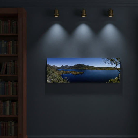 A panoramic landscape wall art print showing a blue lake and Cradle Mountain and sky, displayed on a dark wall under three spotlights next to a bookshelf.