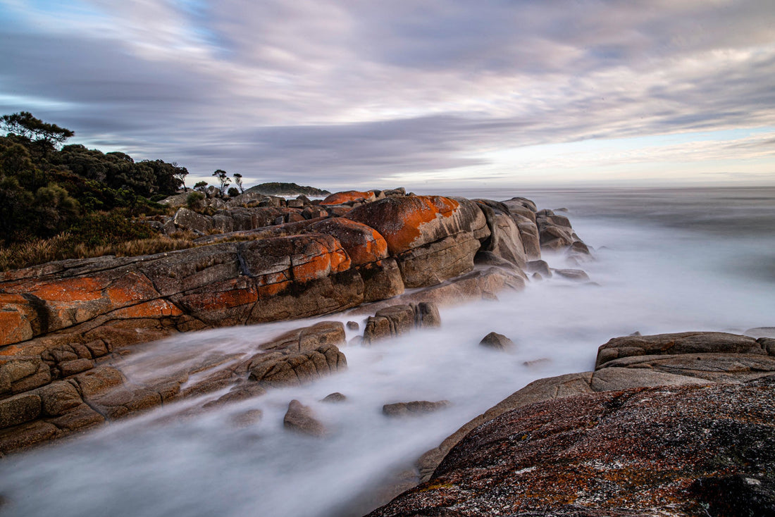 Long exposure of orange lichen-covered granite boulders and misty surf on the Tasmanian coast