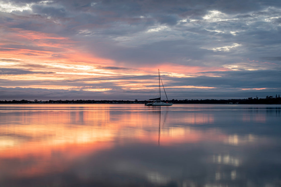 Sailboat silhouetted against a pink and orange sunset reflected on calm water