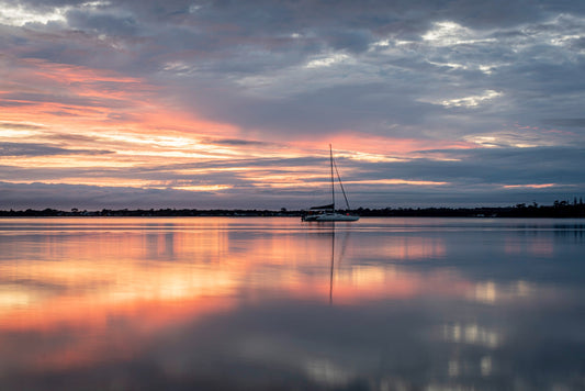 Sailboat silhouetted against a pink and orange sunset reflected on calm water