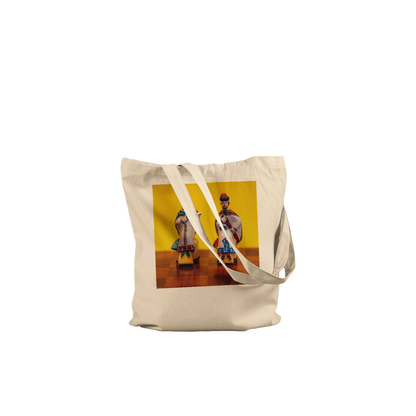 A natural canvas tote bag with a printed graphic of two traditional dancers against a yellow background, shown on a wooden chessboard.