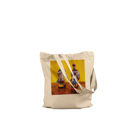 A natural canvas tote bag with a printed graphic of two traditional dancers against a yellow background, shown on a wooden chessboard.