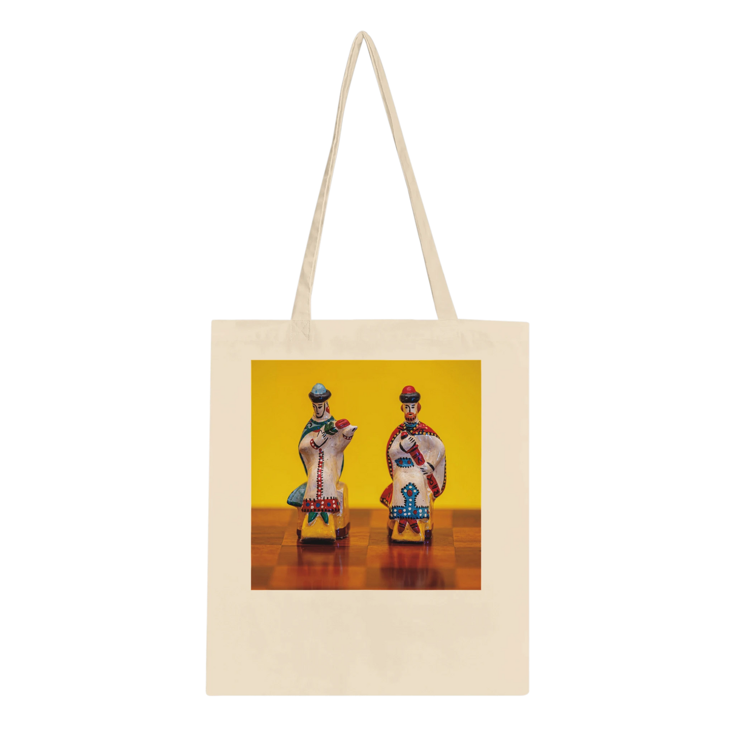 A cream-colored canvas tote bag with long shoulder straps, featuring a square printed graphic on the front. The graphic shows two stylized, colorful figures seated on small chairs against a bright yellow background.