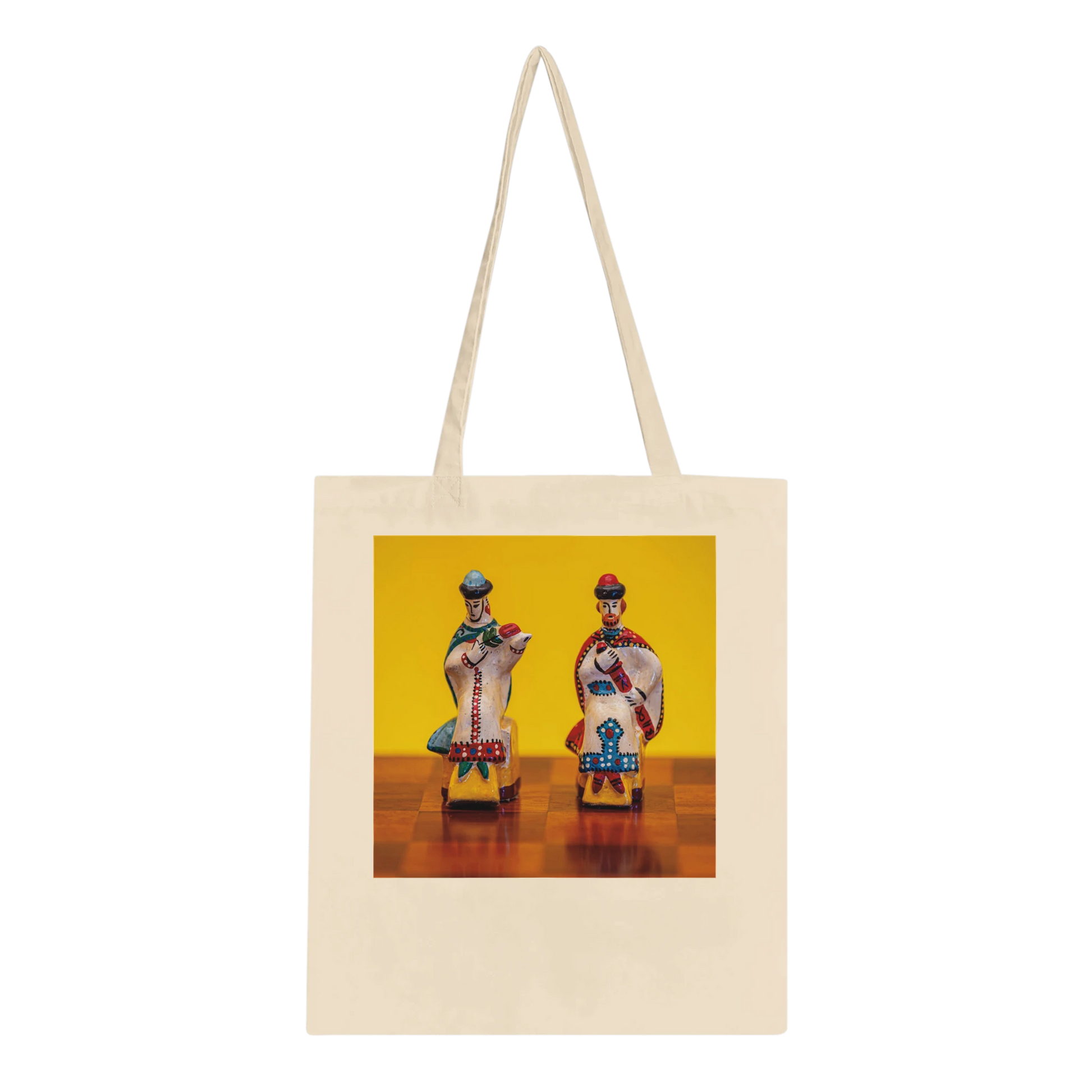 A cream-colored canvas tote bag with long shoulder straps, featuring a square printed graphic on the front. The graphic shows two stylized, colorful figures seated on small chairs against a bright yellow background.