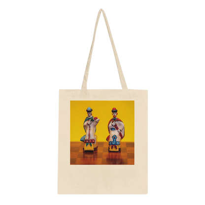 A cream-colored canvas tote bag with long shoulder straps, featuring a square printed graphic on the front. The graphic shows two stylized, colorful figures seated on small chairs against a bright yellow background.