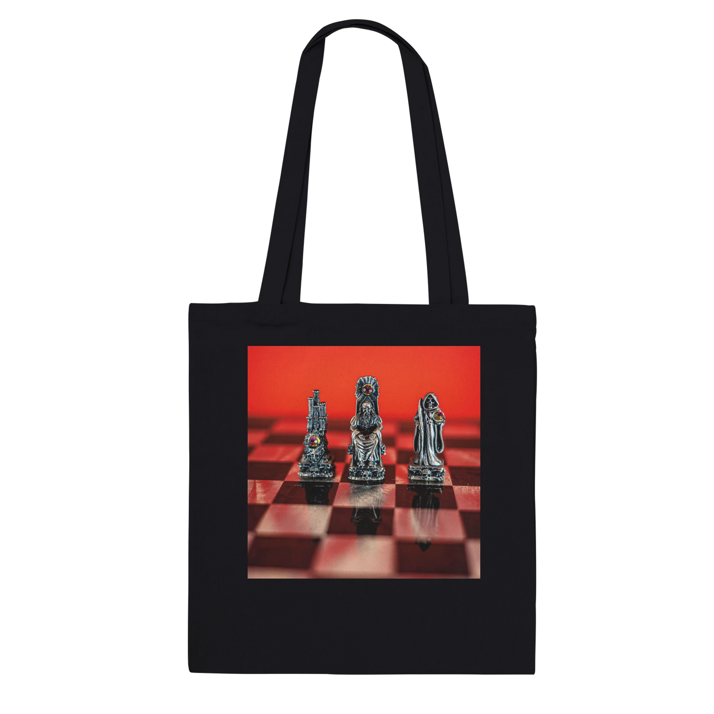 A black canvas tote bag with two shoulder straps, featuring a rectangular printed graphic on the front of three metallic chess pieces on a red and black checkered board against a red background.