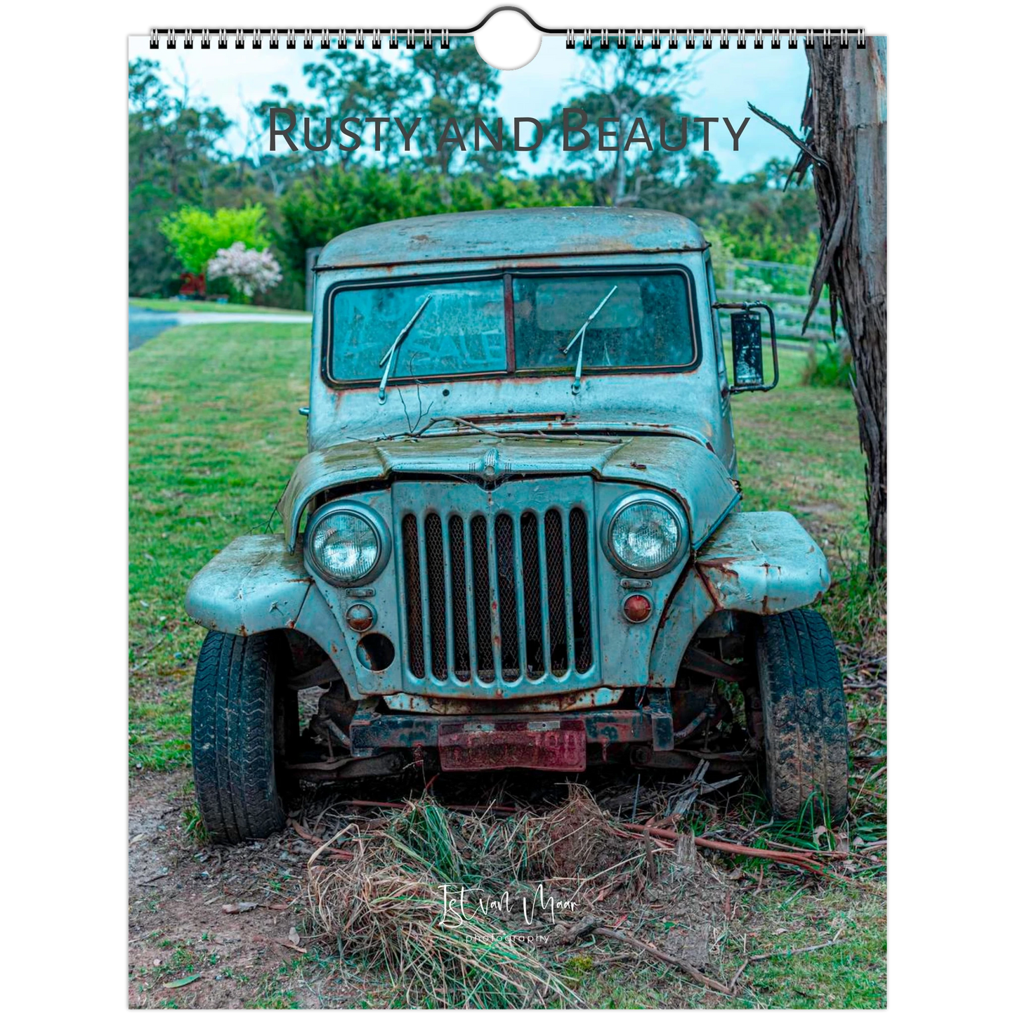 A spiral-bound wall calendar or print featuring an old distressed blue-green jeep parked outdoors on grass and dirt, with the text 'RUSTY AND BEAUTY' across the top. Istvan Maar Photography