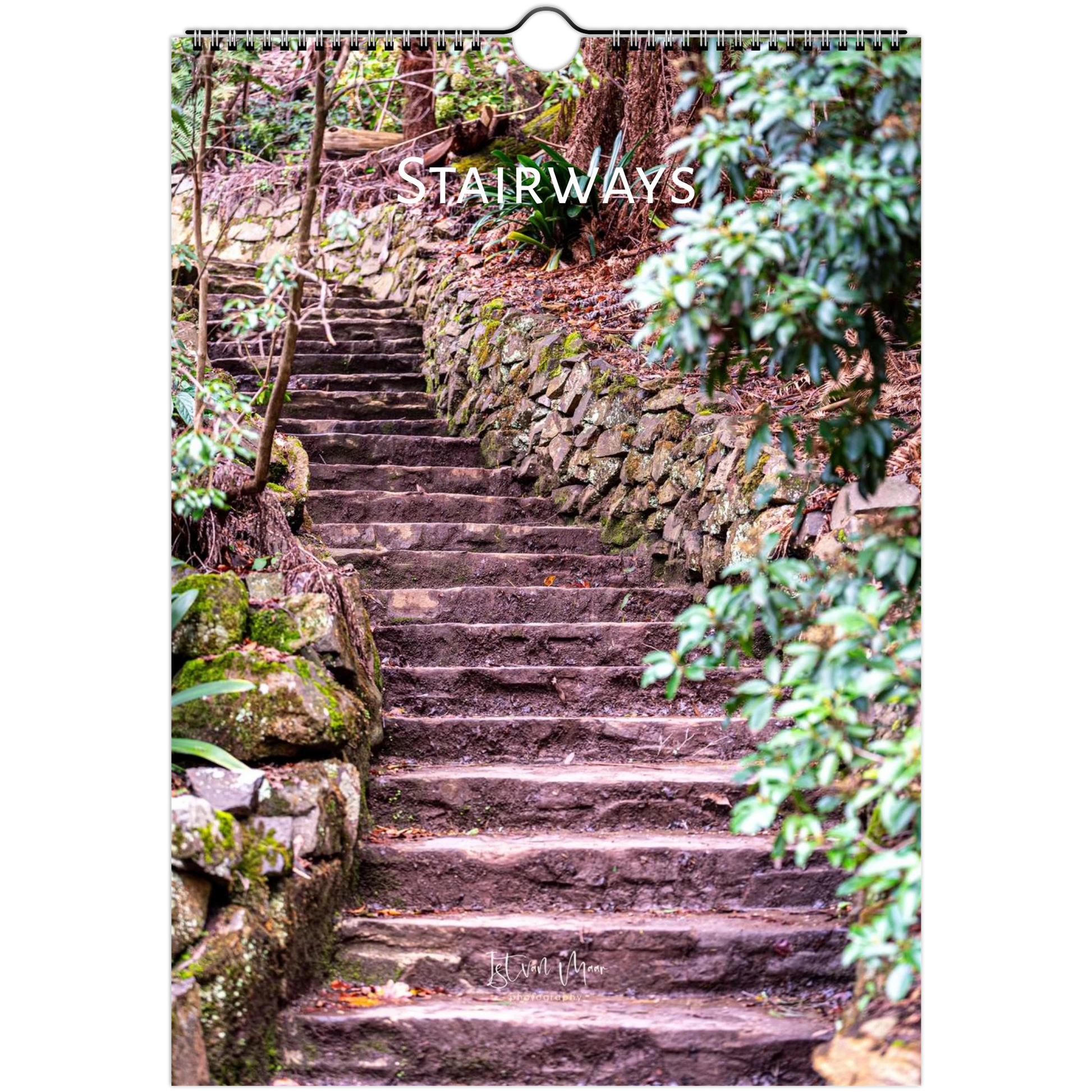 A vertical wall calendar page showing a stone stairway winding through a forest with mossy rocks and green foliage. The word 'STAIRWAYS' appears across the upper middle in white text, and the calendar has a hanging ring at the top.