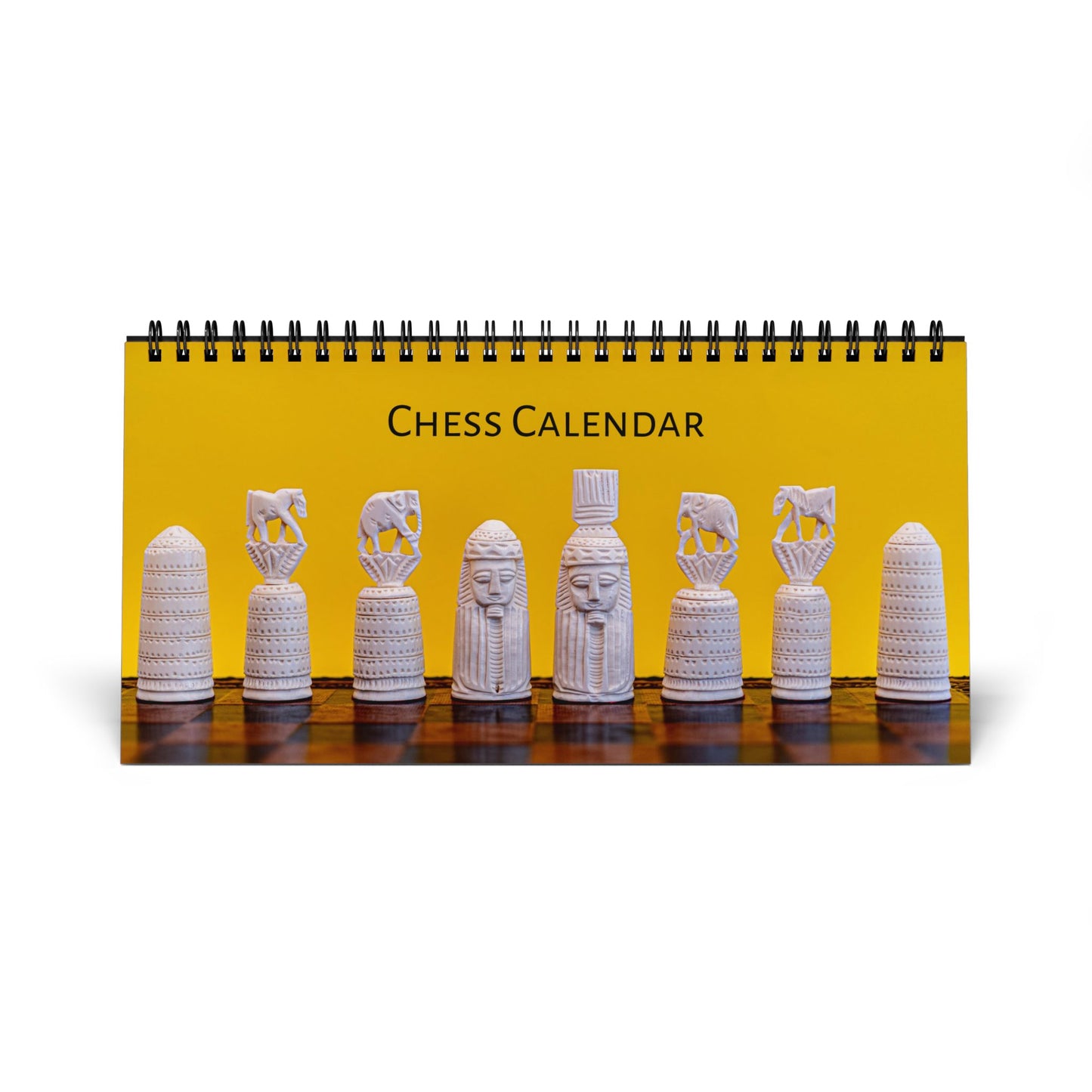 A spiral-bound desk calendar titled 'CHESS CALENDAR' with a bright yellow cover showing a row of white carved chess pieces on a dark wooden surface, photographed on a white background.