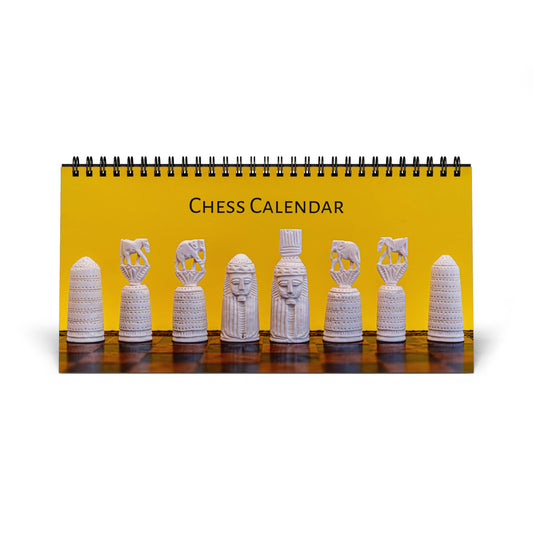 A spiral-bound desk calendar titled 'CHESS CALENDAR' with a bright yellow cover showing a row of white carved chess pieces on a dark wooden surface, photographed on a white background.