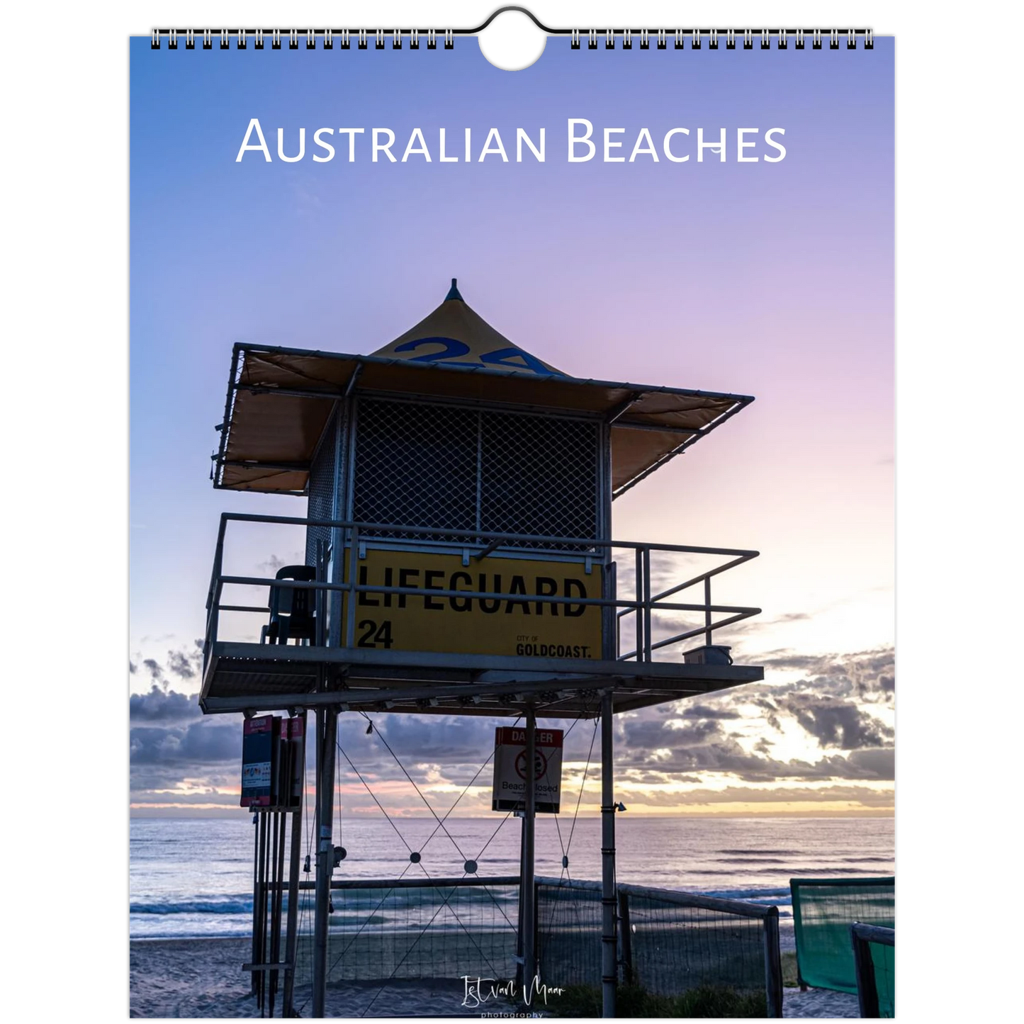 A spiral-bound wall calendar or print titled 'AUSTRALIAN BEACHES' shows a sunset beach scene with a lifeguard tower, ocean, and sky. The product is photographed against a plain white background.