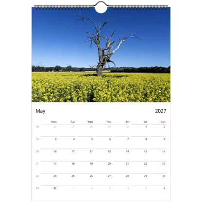 A single-page spiral-bound calendar page showing a landscape photograph of a bare tree in a bright yellow flower field under a clear blue sky, with a May 2027 weekly schedule below.