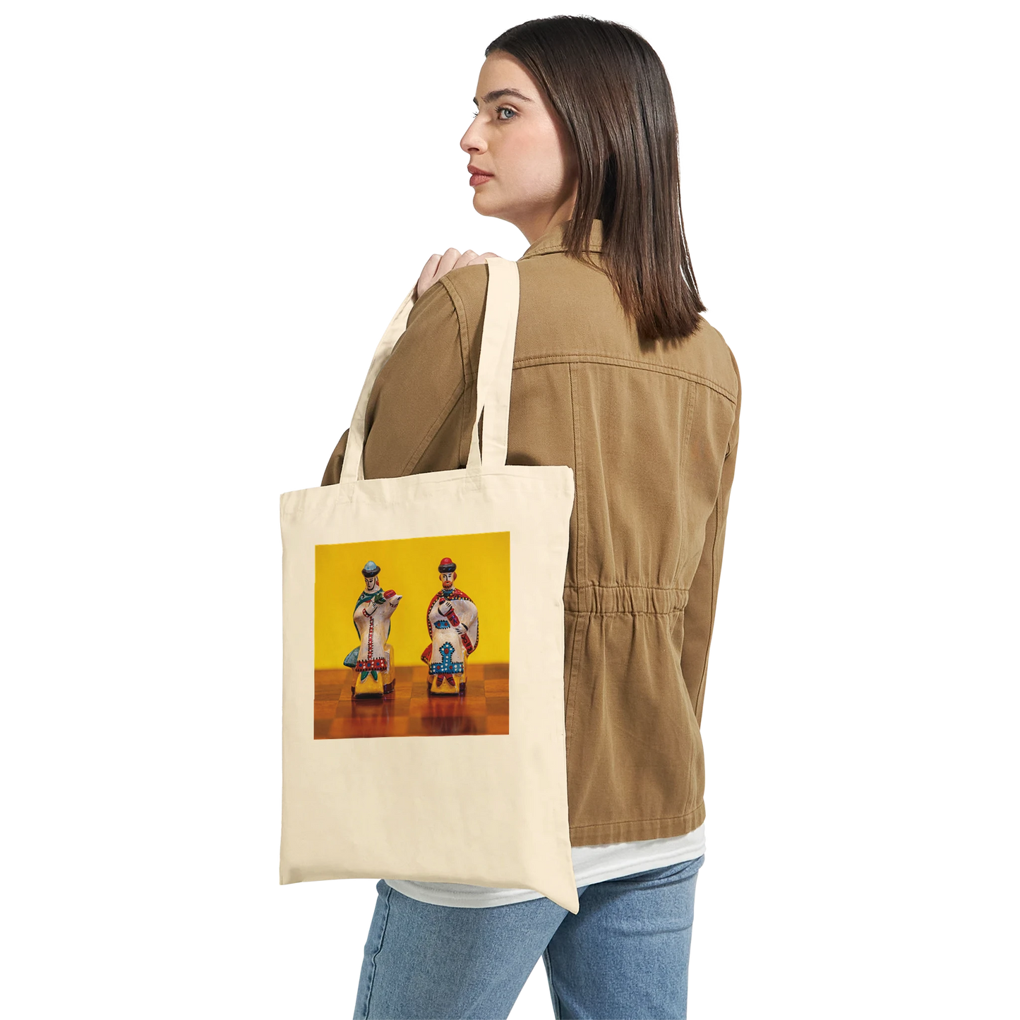 A woman wearing a tan jacket and blue jeans carries a cream canvas tote bag over her shoulder. The tote bag features a colorful graphic print of two stylized figures against a yellow background.