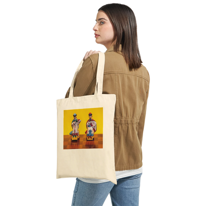 A woman wearing a tan jacket and blue jeans carries a cream canvas tote bag over her shoulder. The tote bag features a colorful graphic print of two stylized figures against a yellow background.