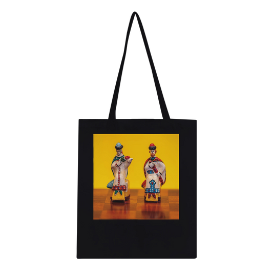 A black canvas tote bag with black shoulder straps, featuring a square graphic print on the front. The print shows two small, colorful traditional figurines standing on a wooden surface against a bright yellow background.