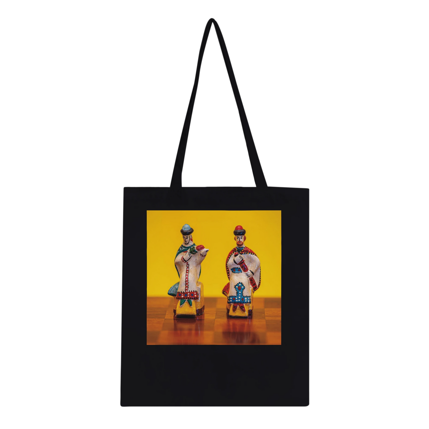 A black canvas tote bag with black shoulder straps, featuring a square graphic print on the front. The print shows two small, colorful traditional figurines standing on a wooden surface against a bright yellow background.
