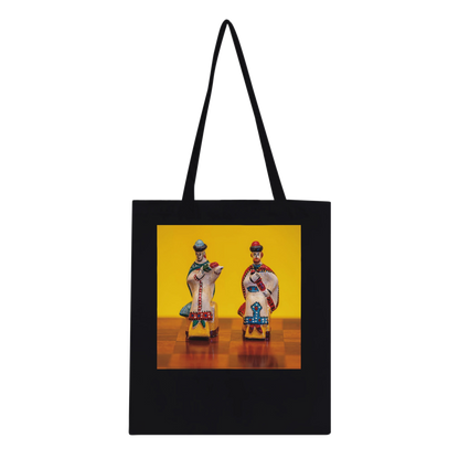 A black canvas tote bag with black shoulder straps, featuring a square graphic print on the front. The print shows two small, colorful traditional figurines standing on a wooden surface against a bright yellow background.