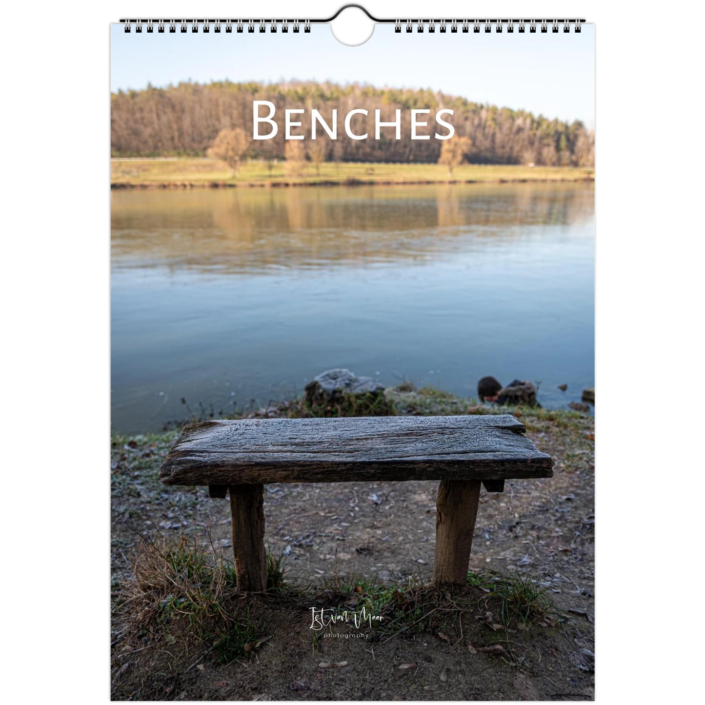 A spiral-bound wall calendar or print titled 'BENCHES' shows a rustic wooden bench in the foreground of a calm lake with trees on the opposite shore. The product is photographed against a plain white background.