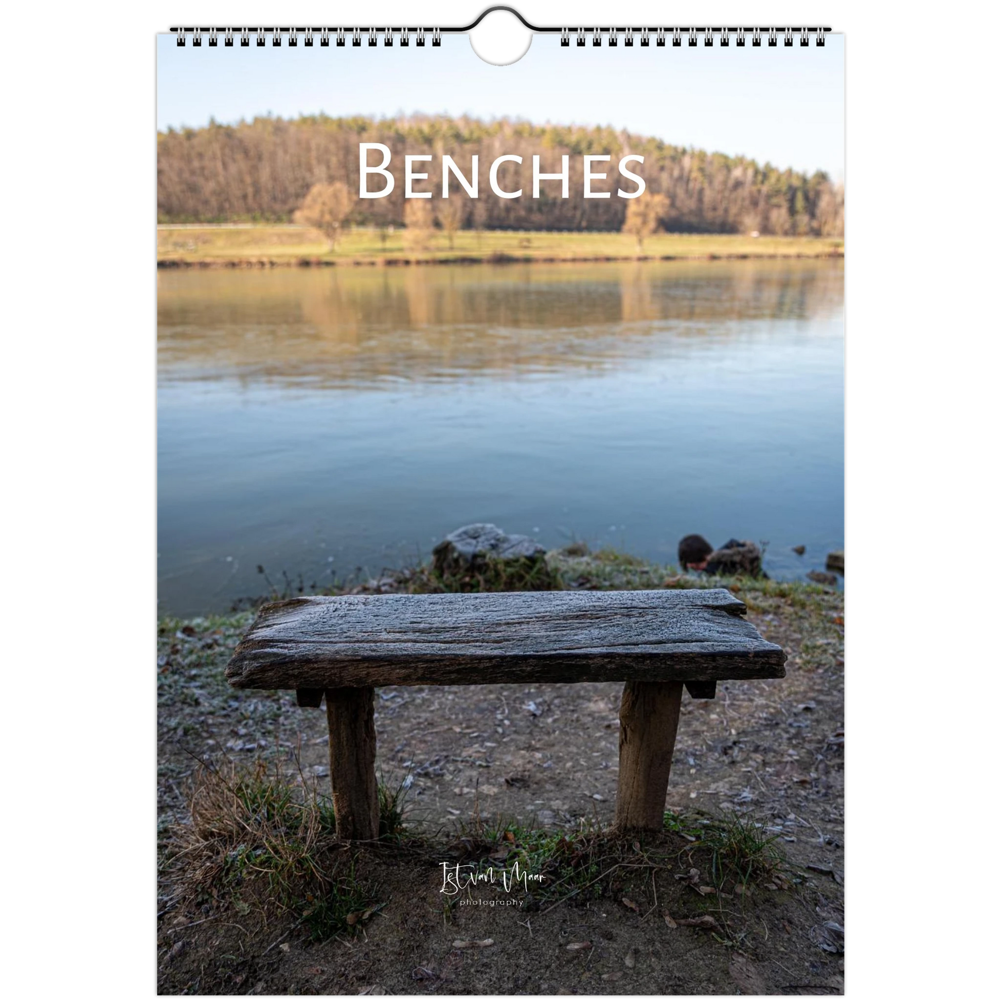 A spiral-bound wall calendar or print titled 'BENCHES' shows a rustic wooden bench in the foreground of a calm lake with trees on the opposite shore. The product is photographed against a plain white background.