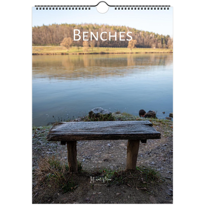 A spiral-bound wall calendar or print titled 'BENCHES' shows a rustic wooden bench in the foreground of a calm lake with trees on the opposite shore. The product is photographed against a plain white background.