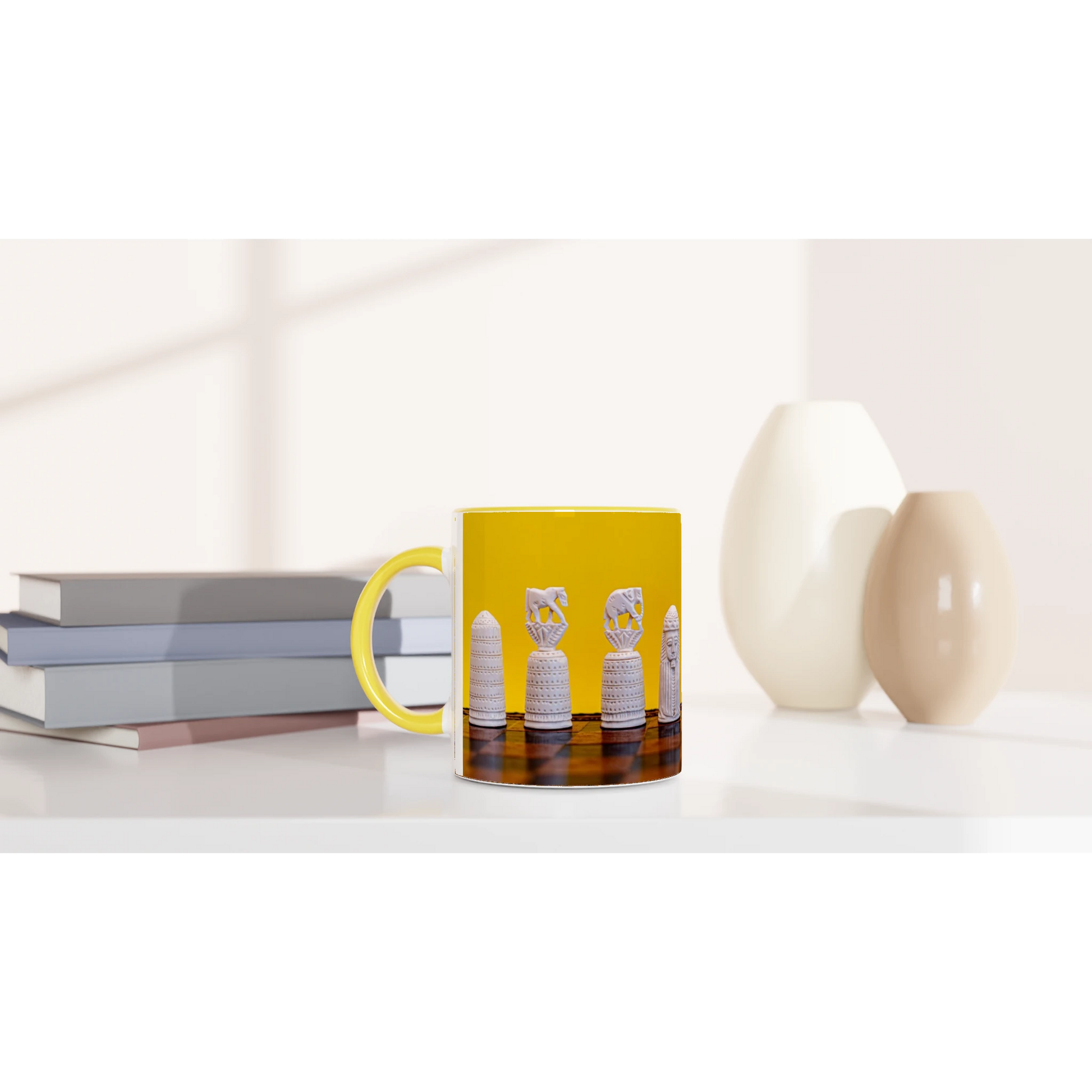 A yellow ceramic mug with a handle, featuring a printed graphic of chess pieces on a dark board, styled on a white surface next to stacked books and two vases.