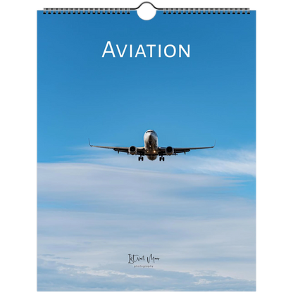 A spiral-bound wall calendar or print titled 'AVIATION' featuring a photograph of an airplane flying through a blue sky with light clouds. The word 'AVIATION' appears in large white letters at the top, and a small signature/brand mark is visible near the bottom.