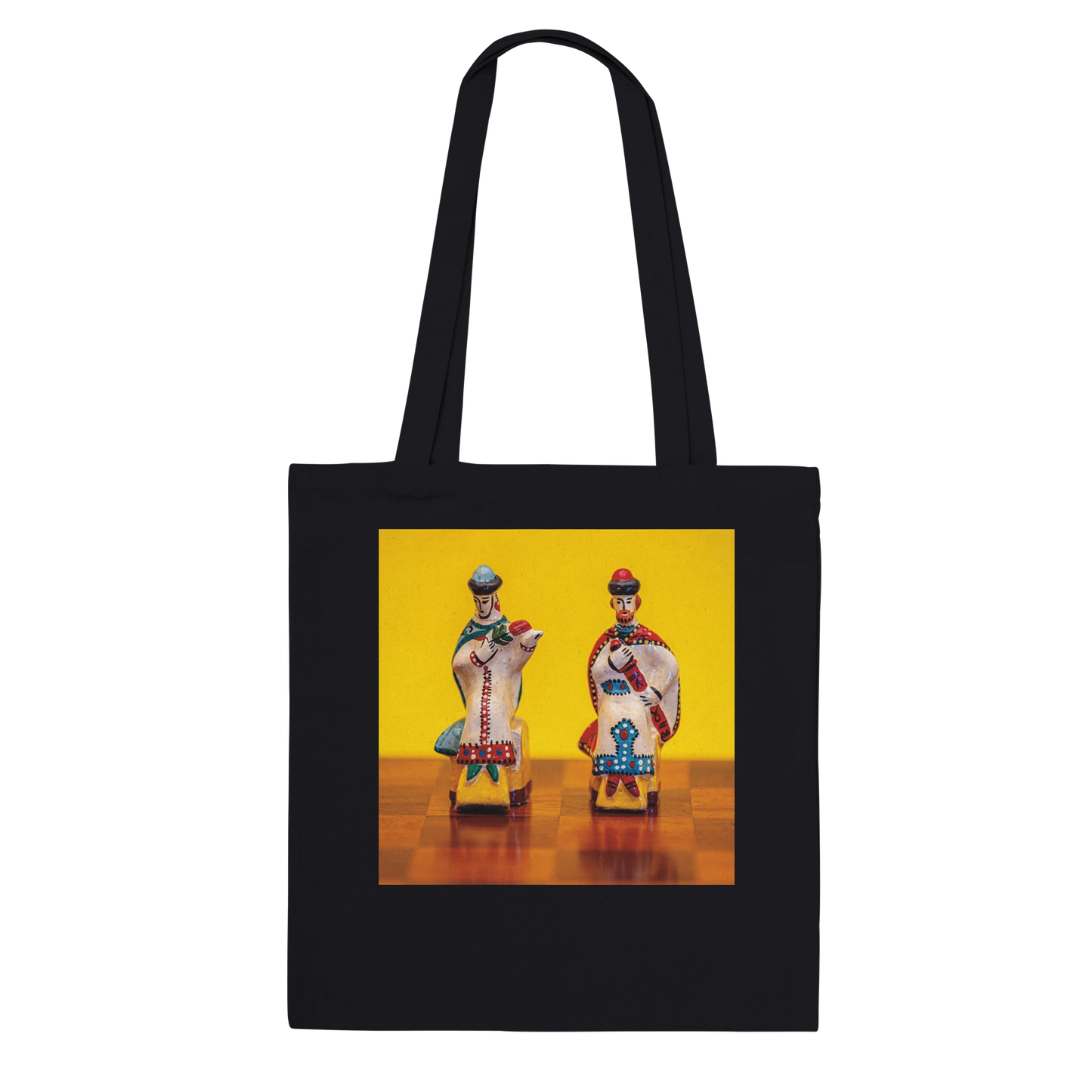 A black canvas tote bag with two shoulder straps, featuring a square graphic print on the front. The print shows two stylized, colorful figures resembling traditional Russian porcelain figurines against a bright yellow background and a brown checkered floor.