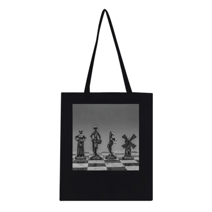 A black canvas tote bag with black shoulder straps, featuring a square black-and-white photographic print of chess pieces on a chessboard across the front. Ships worldwide by Istvan Maar Photography.