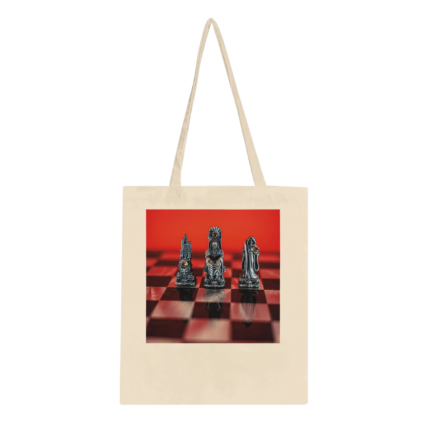 A natural canvas tote bag with long handles, featuring a rectangular printed graphic on the front of three metallic chess pieces on a red and black checkered board against a solid red background.