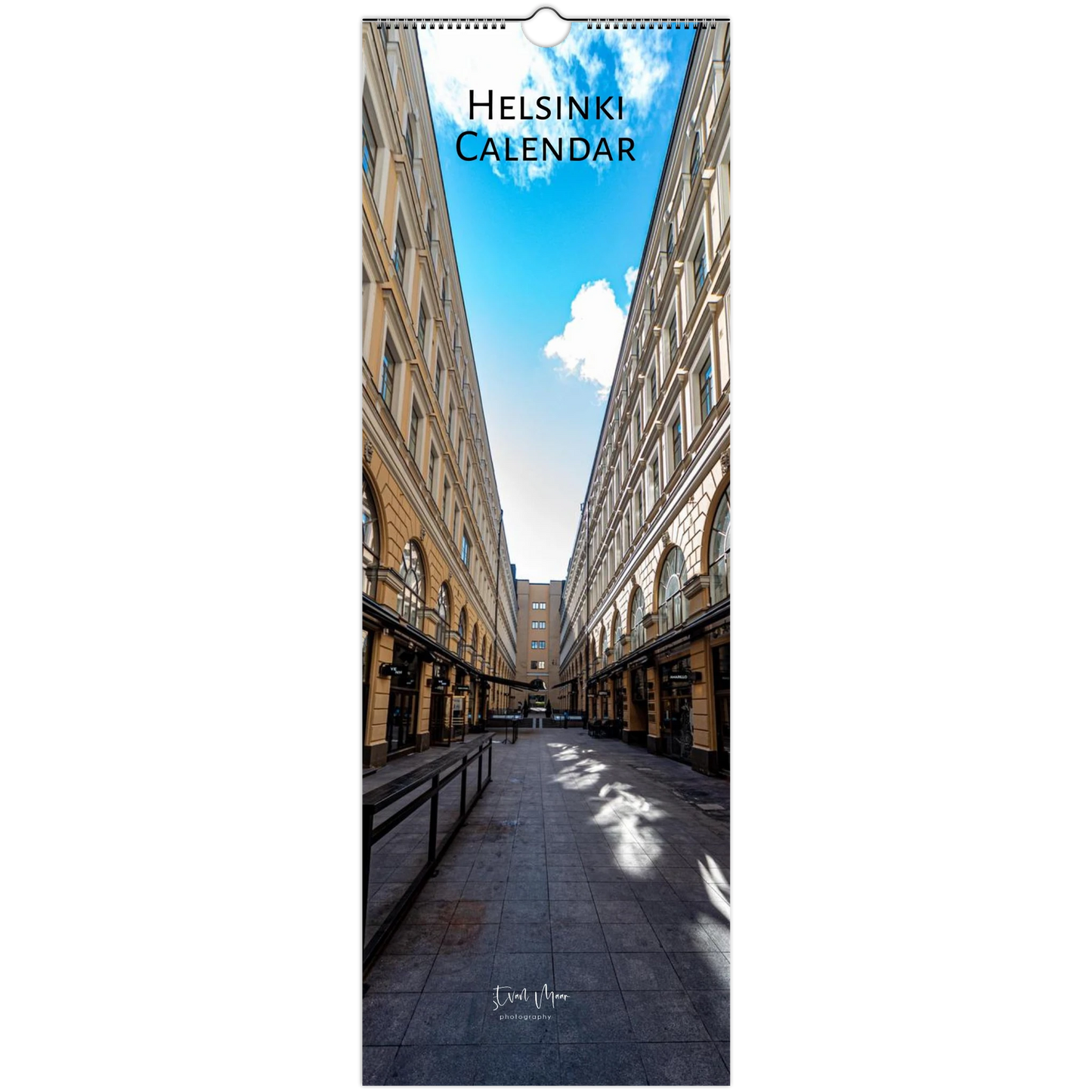 A vertical wall calendar titled 'HELSINKI CALENDAR' showing a photograph of a city street flanked by tall historic buildings under a blue sky with clouds. The calendar has a hanging tab at the top and is photographed against a plain white background.