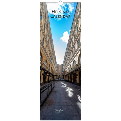 A vertical wall calendar titled 'HELSINKI CALENDAR' showing a photograph of a city street flanked by tall historic buildings under a blue sky with clouds. The calendar has a hanging tab at the top and is photographed against a plain white background.