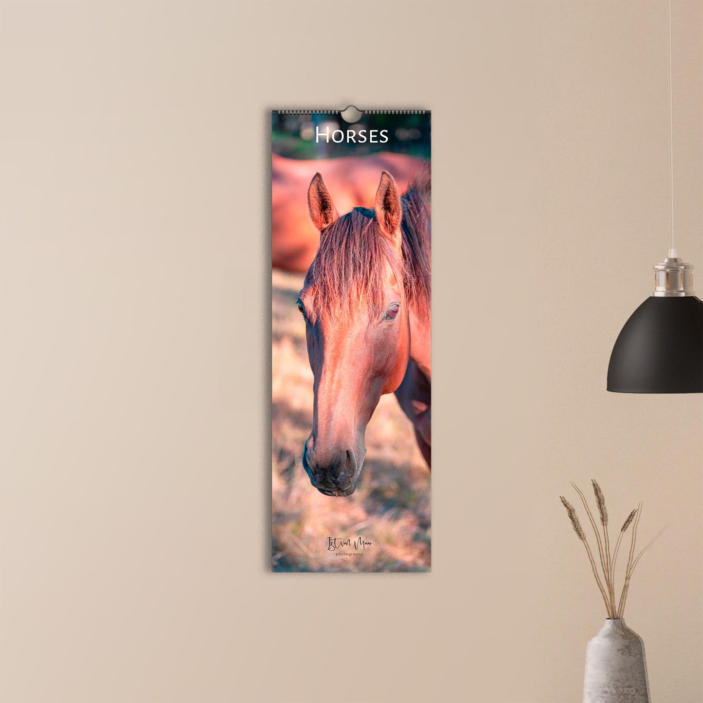 A vertical slim wall calendar featuring a close-up photograph of a horse's face, hanging on a beige wall in a styled interior setting with a black pendant lamp and a vase. 