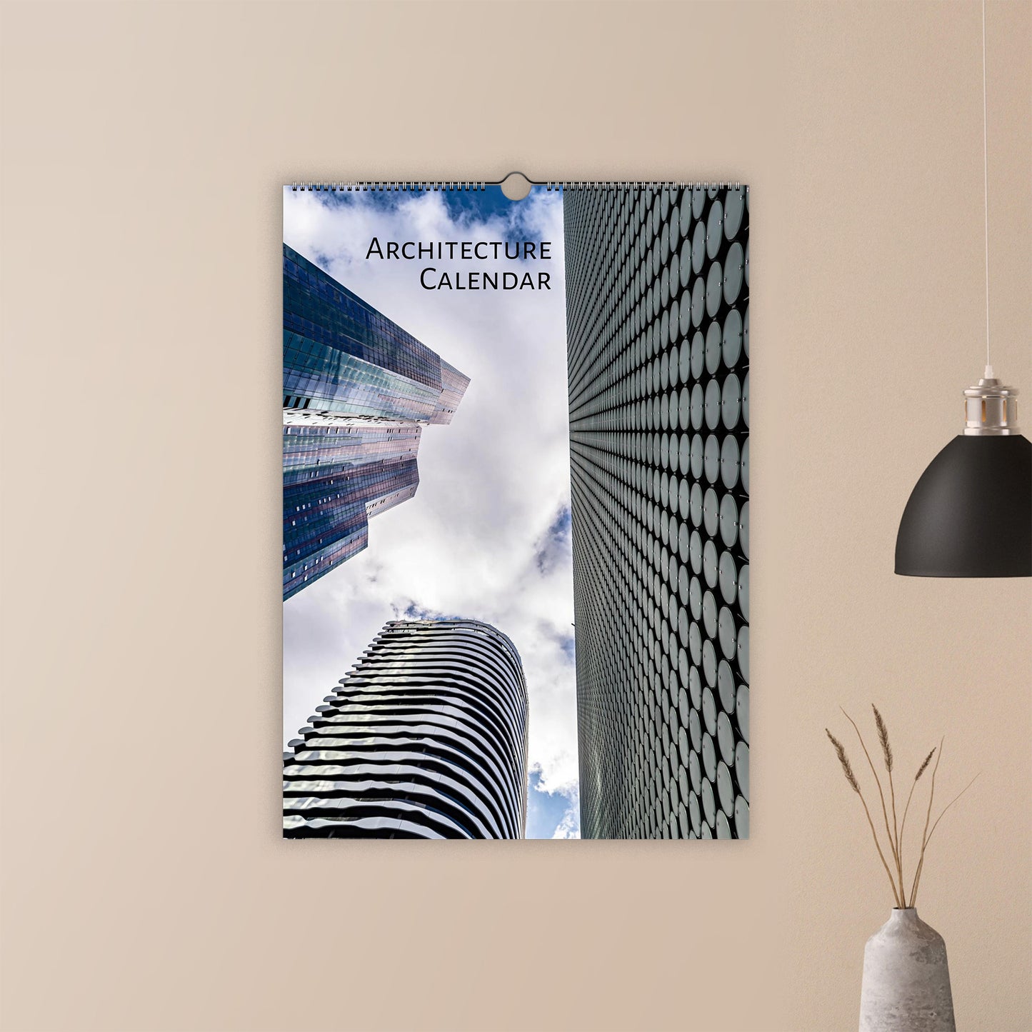 A spiral-bound wall calendar titled 'Architecture Calendar' hanging on a beige wall. The calendar cover features a striking architectural photograph of modern skyscrapers, one with a black-and-white circular pattern and another looking up at the sky.