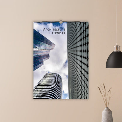 A spiral-bound wall calendar titled 'Architecture Calendar' hanging on a beige wall. The calendar cover features a striking architectural photograph of modern skyscrapers, one with a black-and-white circular pattern and another looking up at the sky.