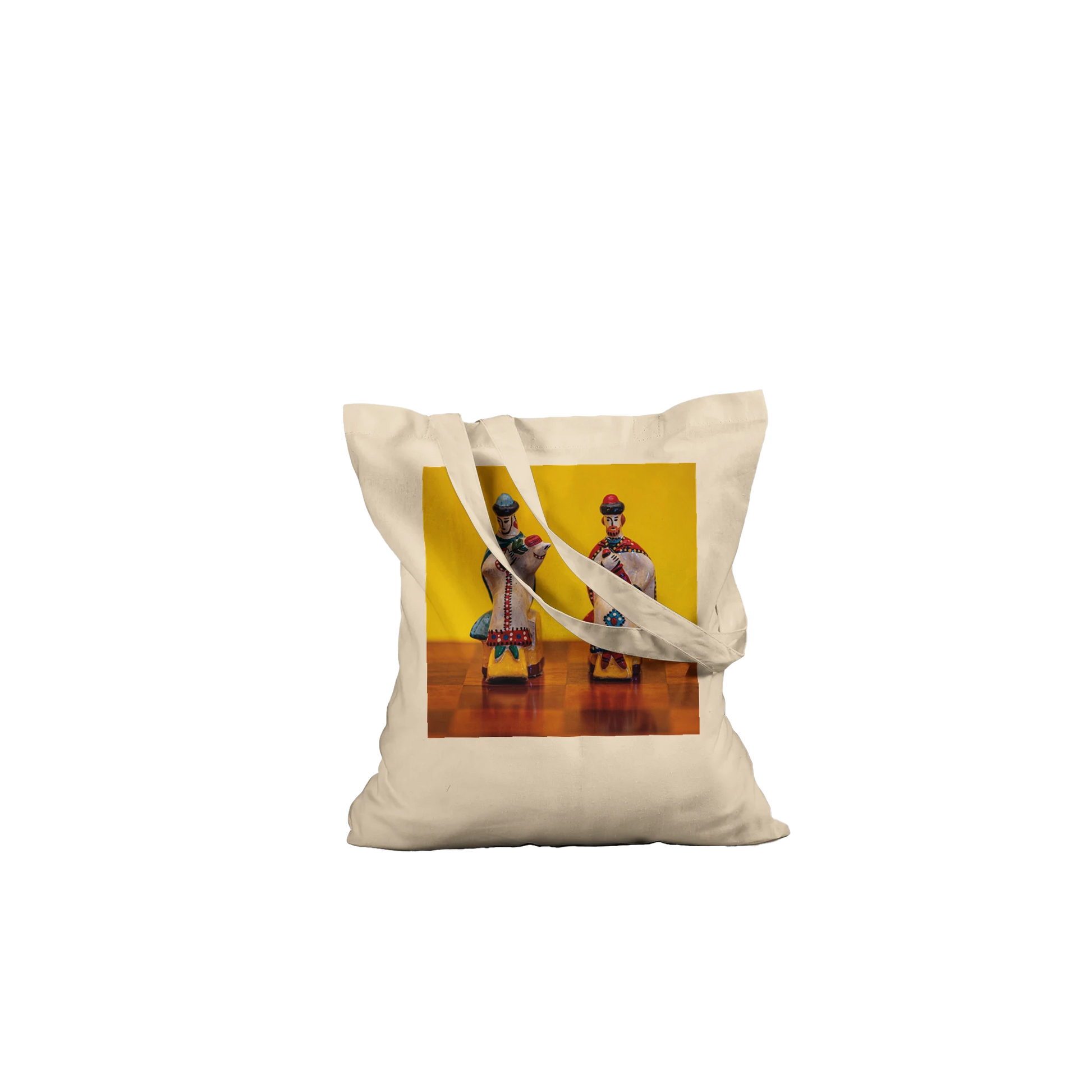 A natural canvas tote bag with two shoulder straps, featuring a square printed graphic on the front. The graphic shows two traditional Russian porcelain figurines against a bright yellow background.