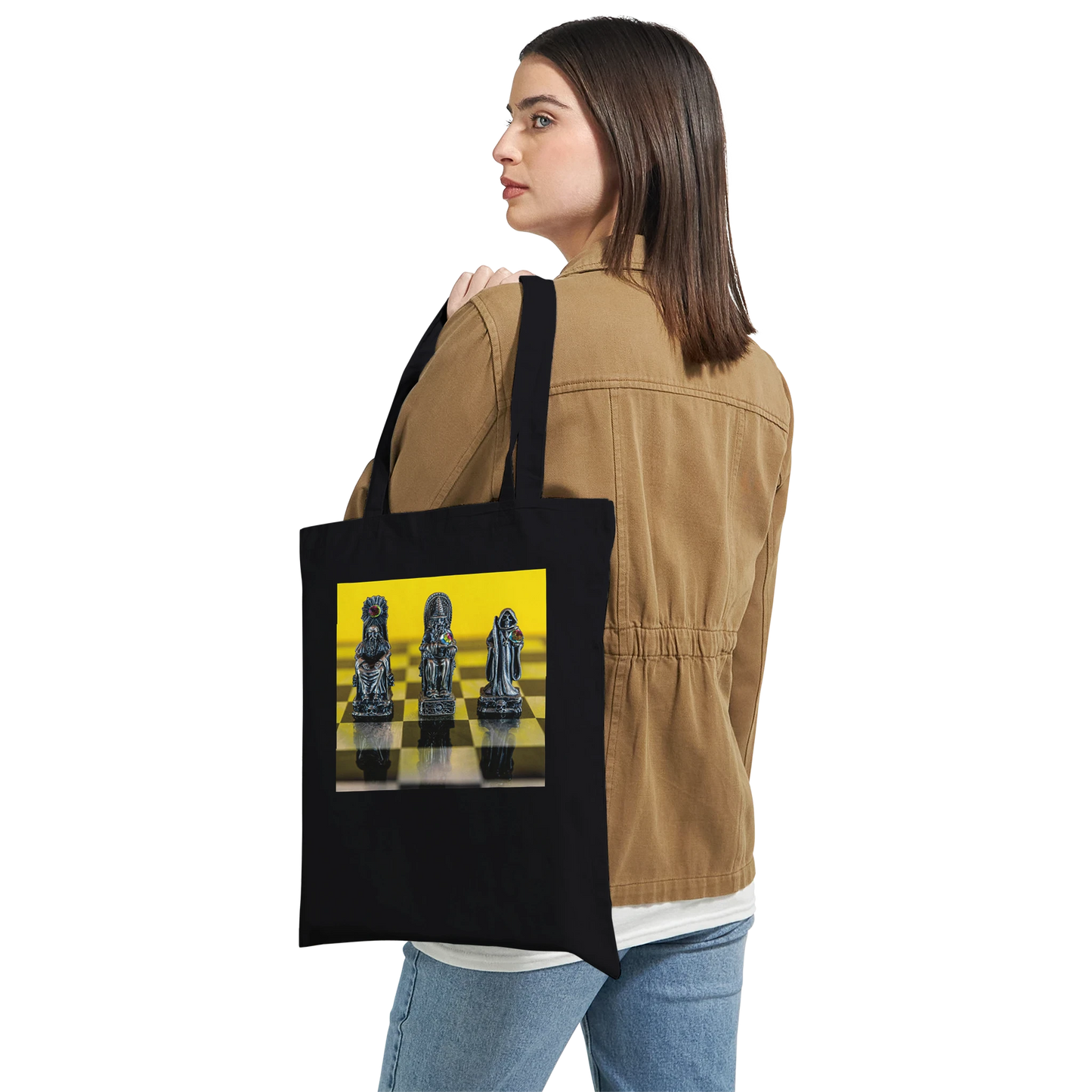 A woman wearing a tan jacket and blue jeans is shown from the back, carrying a black canvas tote bag over her shoulder. The tote bag features a graphic print of chess pieces on a yellow and black checkered board.