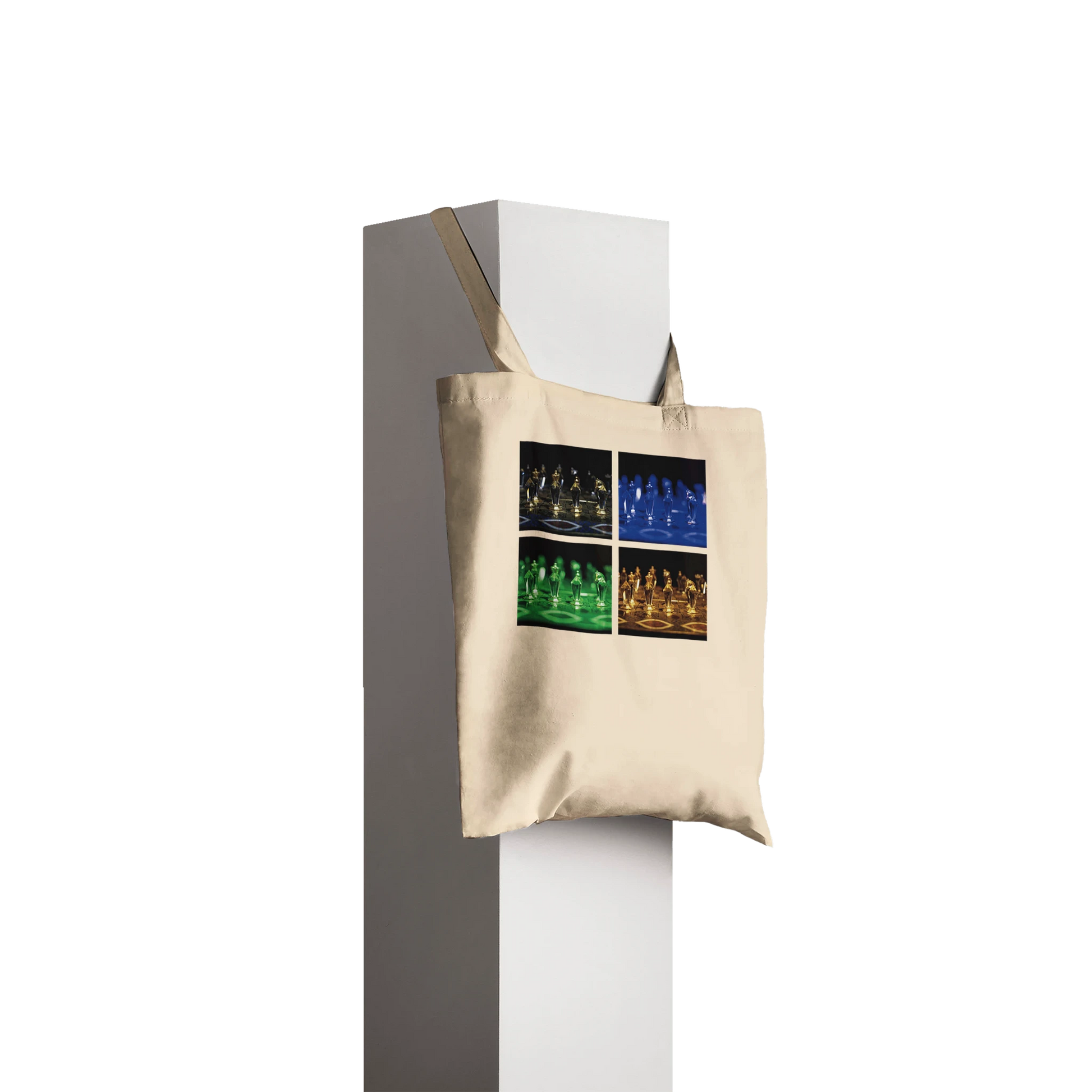 A beige canvas tote bag with a four-panel pop-art style graphic of chess pieces in black, blue, green, and gold, hanging from a white display stand against a dark gray background.