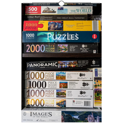 A tall retail display or shelf filled with boxed jigsaw puzzles from multiple brands, including Arthur Boyd, The Great Hall Tapestry, Woodland, Panoramic, and Images of the World. The boxes are stacked horizontally, showing different puzzle types and piece counts. 2027 Puzzle Wall Calendar – front cover featuring puzzle photography by Istvan Maar.