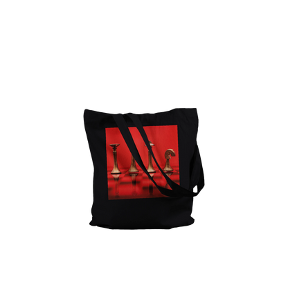 A black canvas tote bag with black shoulder straps, featuring a square graphic print on the front depicting metallic gold chess pieces on a vibrant red background.
