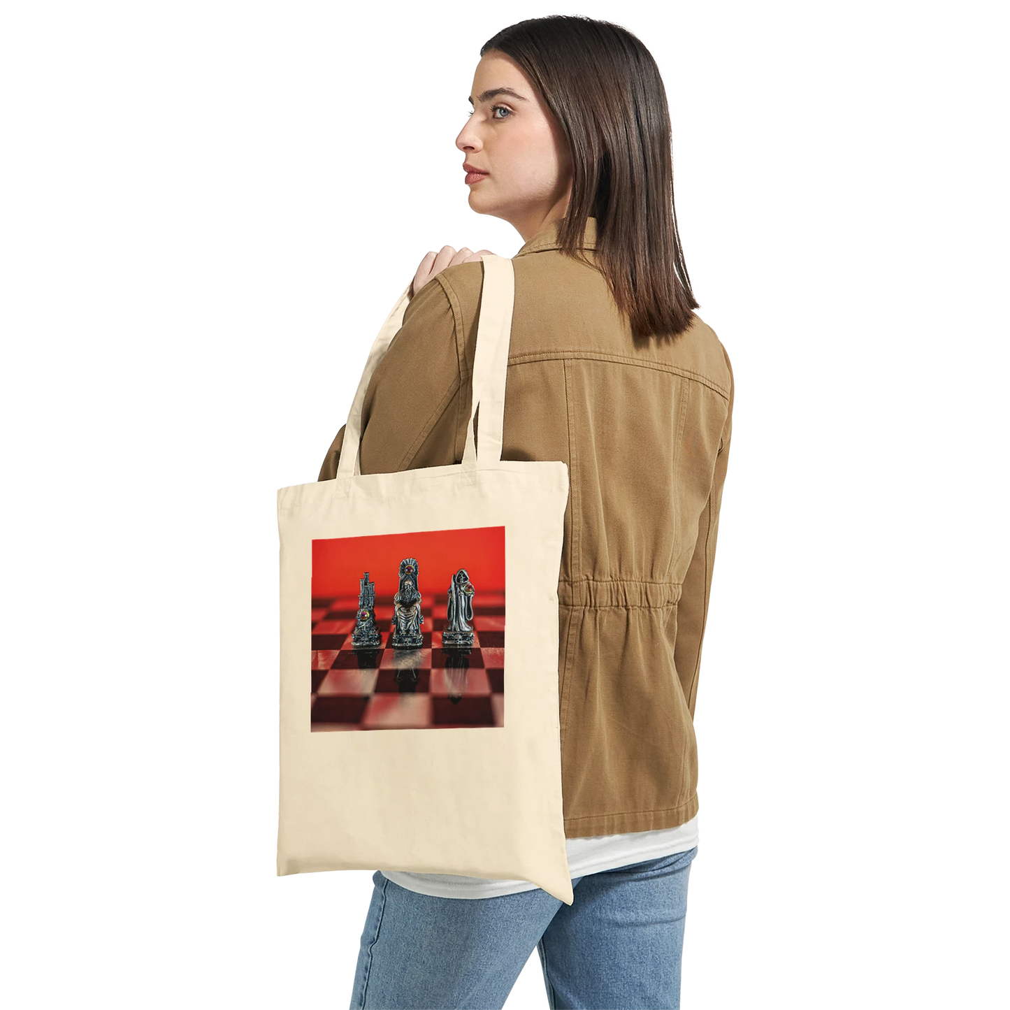 A woman wearing a tan jacket and blue jeans is shown from the back holding a cream canvas tote bag over her shoulder. The tote bag features a printed graphic of black chess pieces on a red and black checkered board.