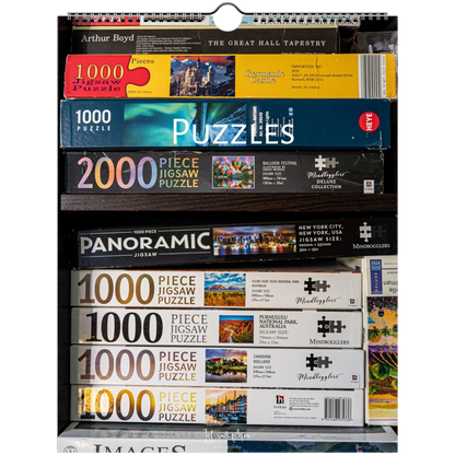 A retail display shelf filled with boxed jigsaw puzzles from multiple brands, including Arthur Boyd, Mindboggle, and Monopoly. The boxes are stacked by size, with 1000-piece and 2000-piece puzzles visible.