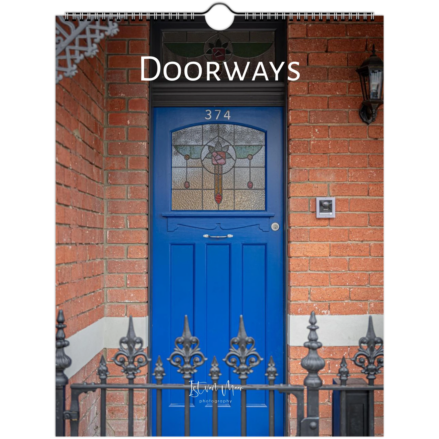 A spiral-bound wall calendar page titled 'Doorways' featuring a photograph of a bright blue front door set in a red brick wall, with decorative iron railings and the house number 374.