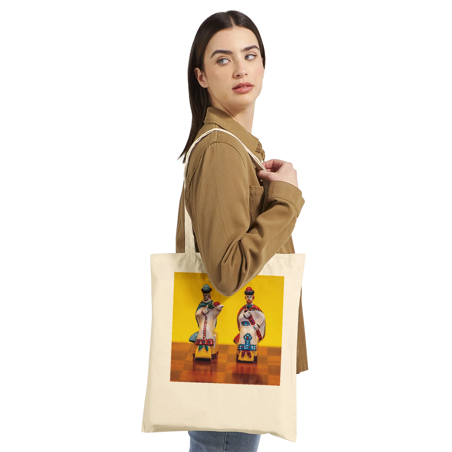 A woman wearing a brown jacket carries a cream canvas tote bag with a colorful graphic print of two stylized figures on a yellow background. The photo is shot against a solid white studio background.