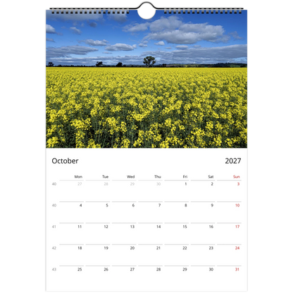 A spiral-bound wall calendar page showing a photograph of a bright yellow flower field under a blue sky with clouds, with the calendar grid for October 2027 below.