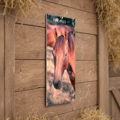 A wall calendar featuring a close-up photograph of a brown horse, hanging on a rustic wooden barn door with hay visible in the corners.