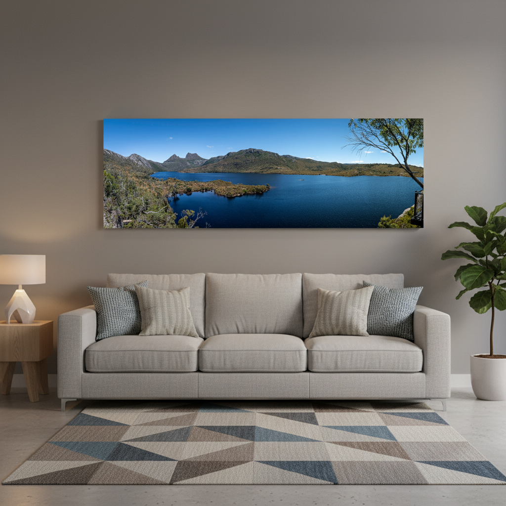A panoramic landscape wall art print showing a blue lake, Cradle Mountain, and sky, displayed above a light beige sofa in a modern living room setting.