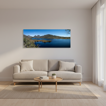 A panoramic canvas print of a scenic Cradle Mountain lake landscape is displayed on a light beige wall above a modern light gray sofa in a bright, minimalist living room.