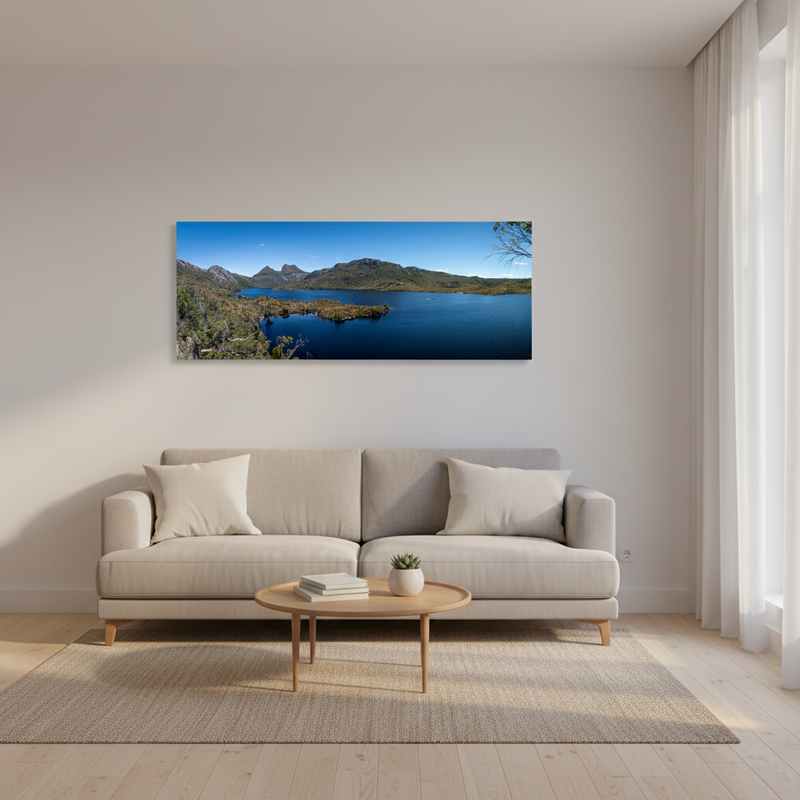 A panoramic canvas print of a scenic Cradle Mountain lake landscape is displayed on a light beige wall above a modern light gray sofa in a bright, minimalist living room.