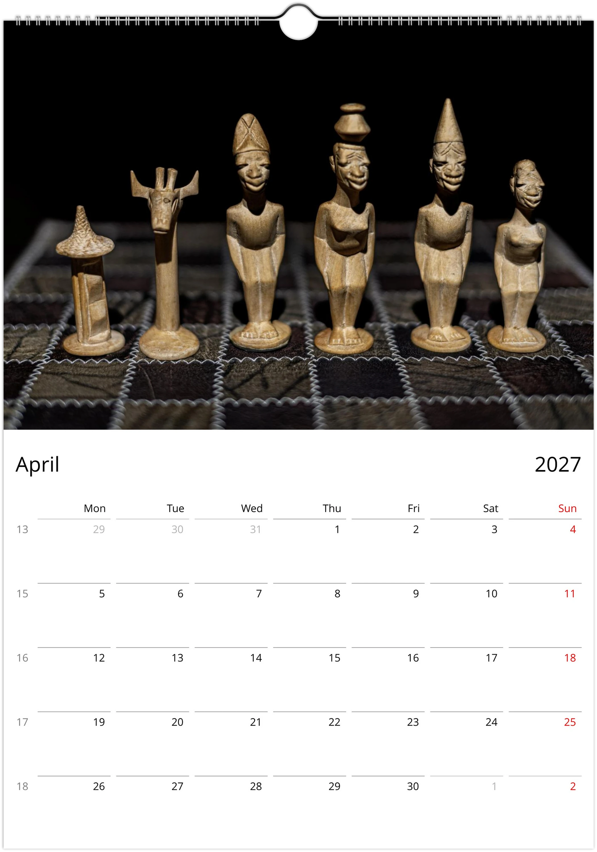 A wall calendar page showing the month of April 2027 below a large photograph of six carved wooden figurines on a dark grid surface. The calendar has a white lower section with black text and a small red day highlight for the 4th.